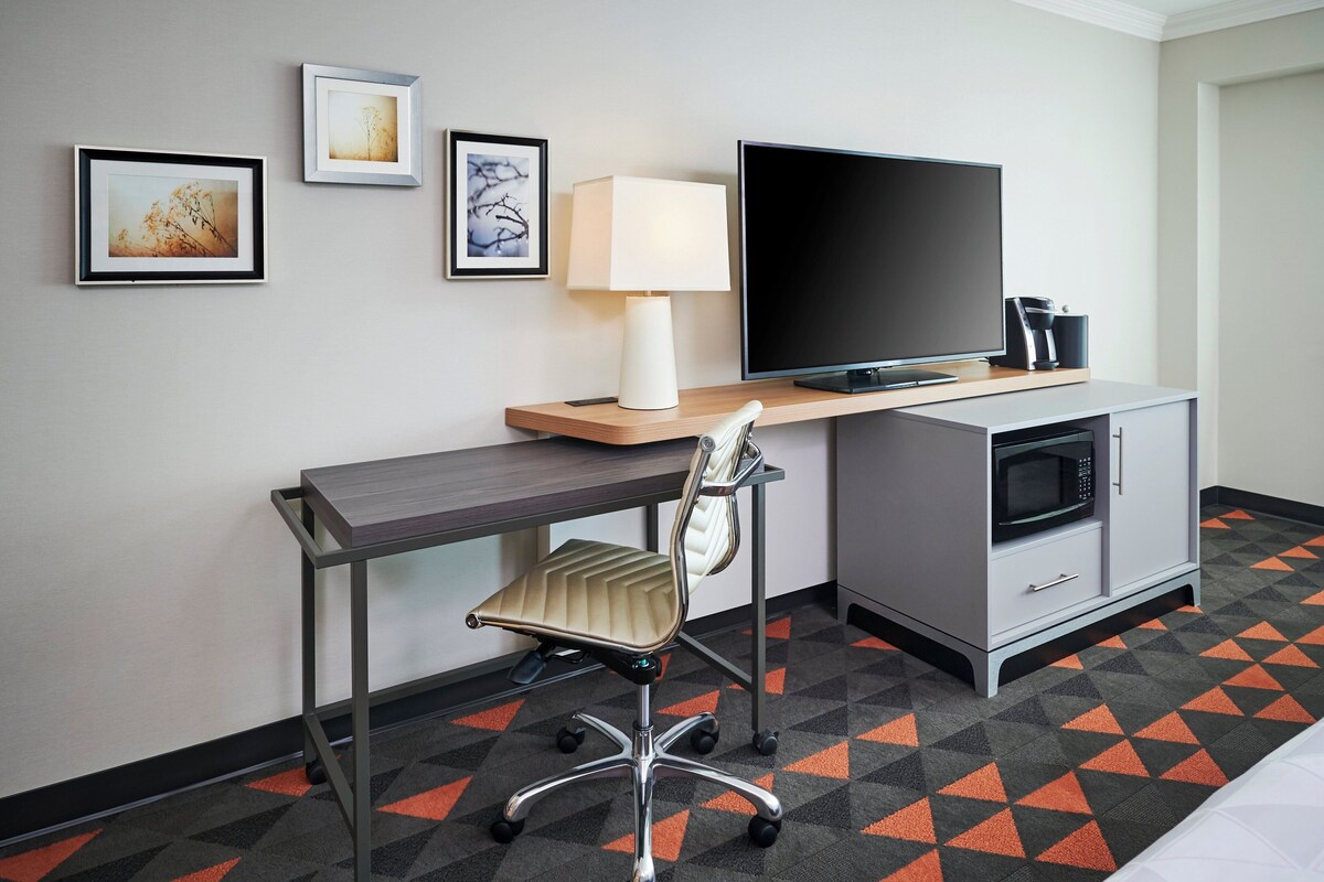 A hotel room with a desk, chair, television, lamp, microwave, and a cabinet.