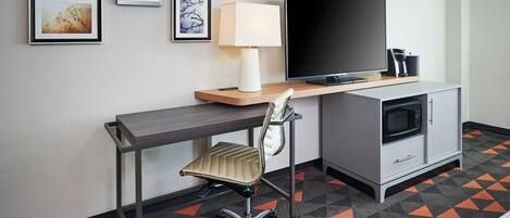 Habitación de hotel con escritorio, silla, televisor, lámpara, microondas y un armario.