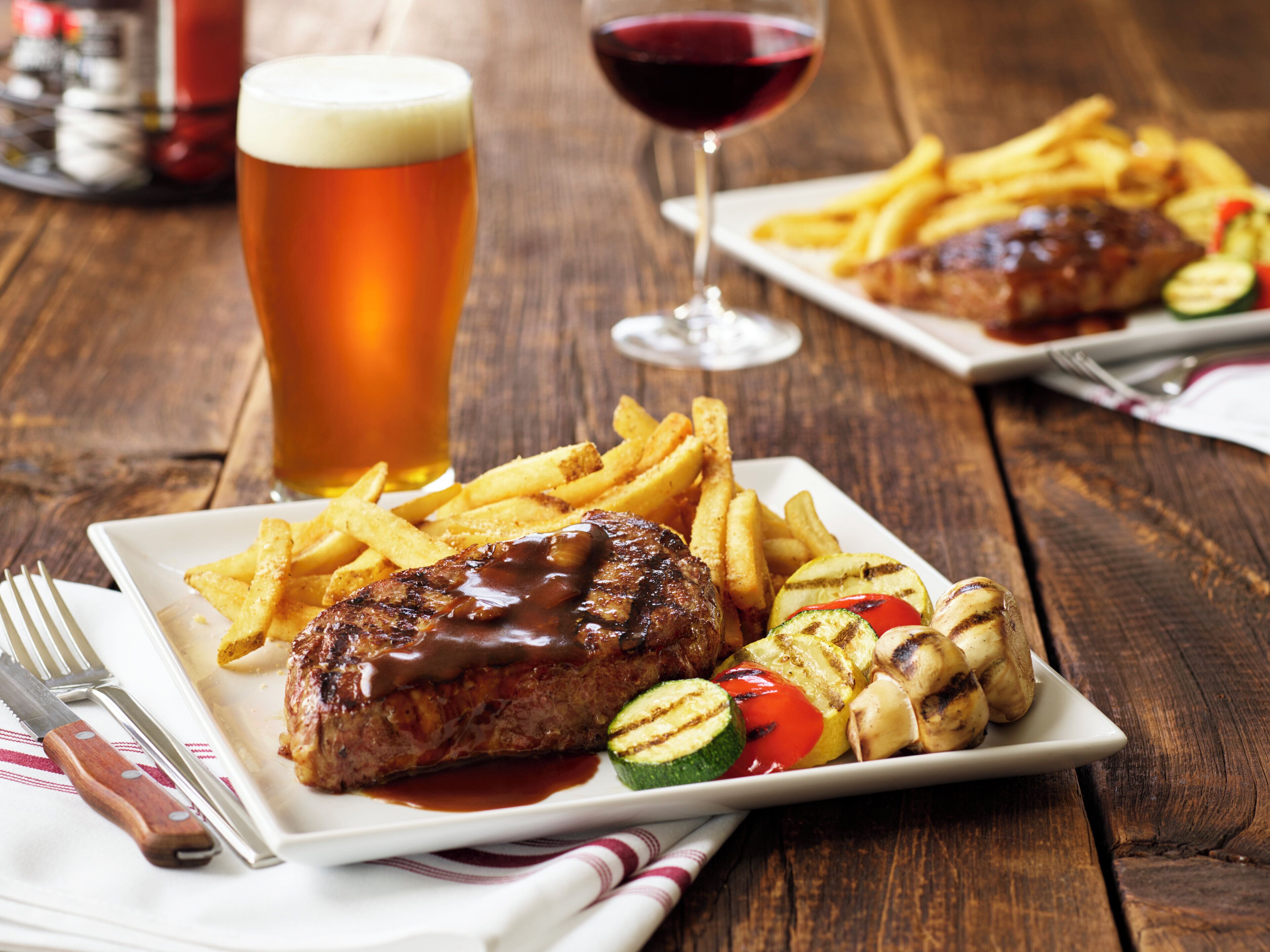Een bord eten met een steak, frietjes en gegrilde groenten, vergezeld van een glas bier en een glas rode wijn.