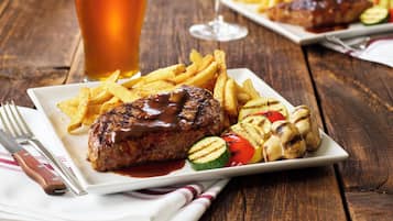 Een bord eten met een steak, frietjes en gegrilde groenten, vergezeld van een glas bier en een glas rode wijn.