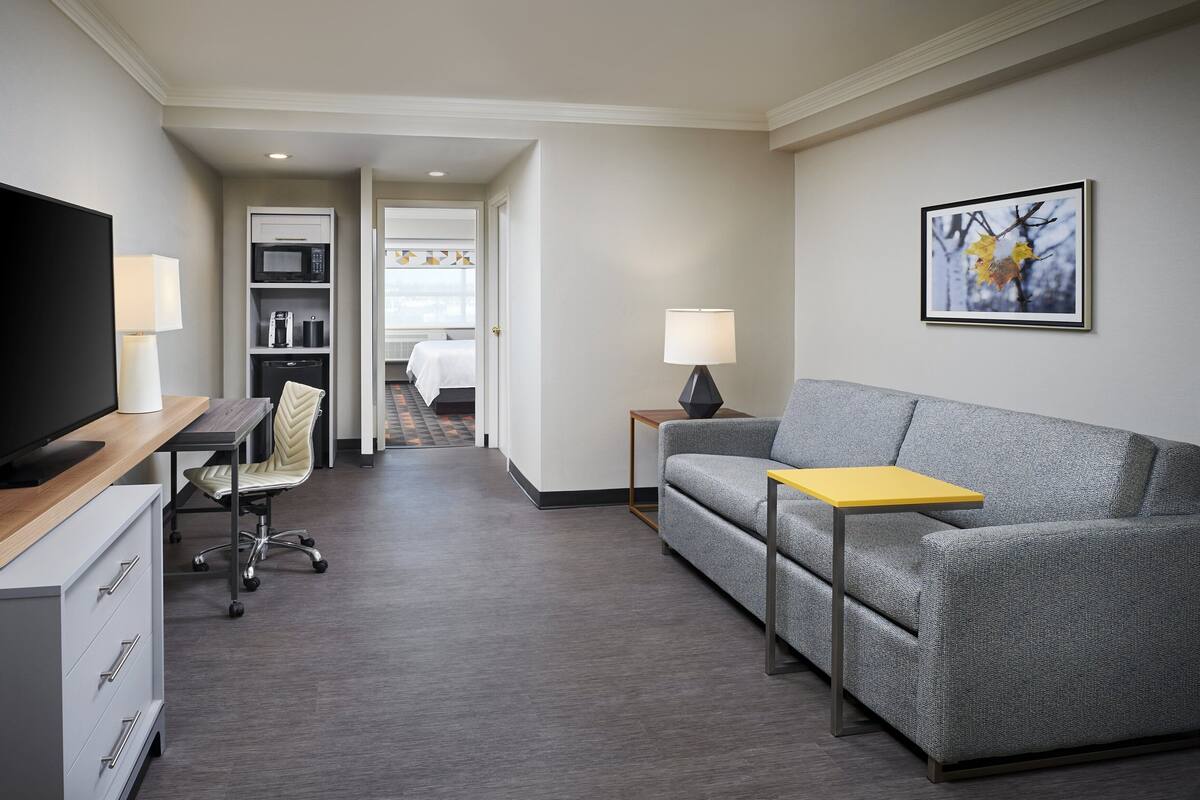 A modern hotel room with a flat-screen TV, a sofa, a small table, a desk with a lamp, a chair, a microwave, and a bed.