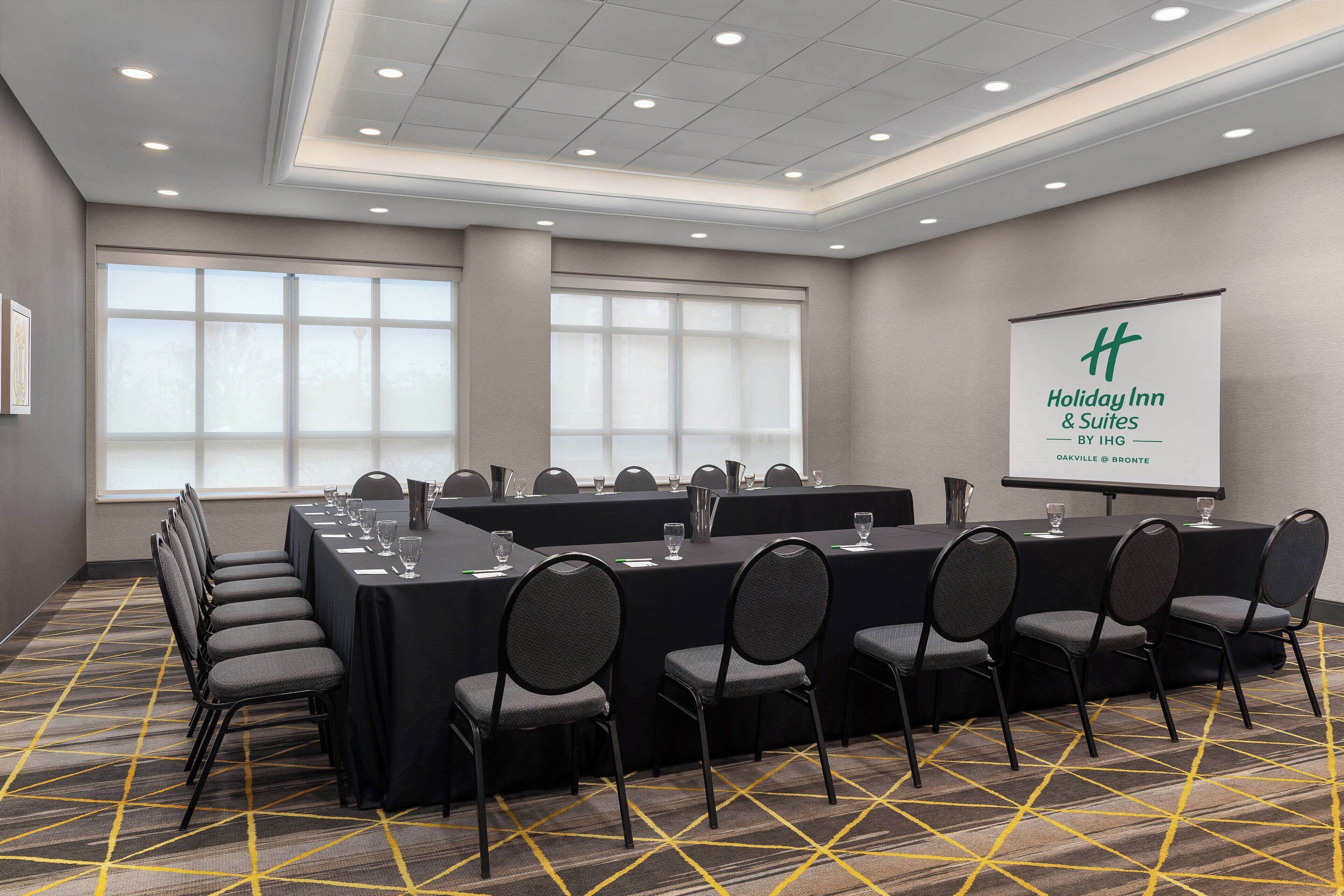 Une salle de conférence équipée d’une longue table, de chaises et d’un écran de projection affichant le nom « Holiday Inn & Suites by IHG Oakville & Bronte ».