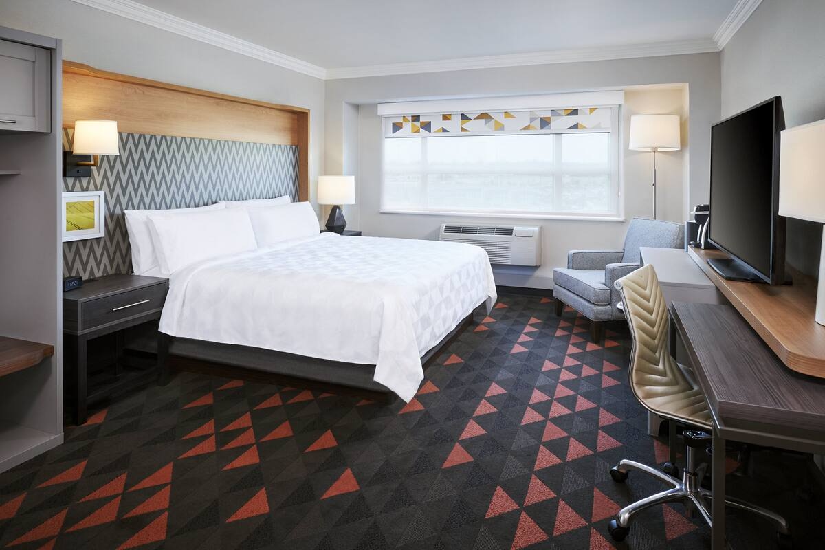 A hotel room with a bed, a desk with a chair, a TV, and a window with a view.