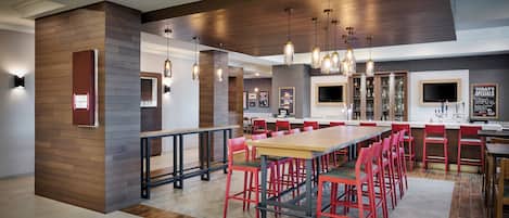 A modern restaurant interior with a long communal table, red chairs, pendant lighting, and a bar area with a television.