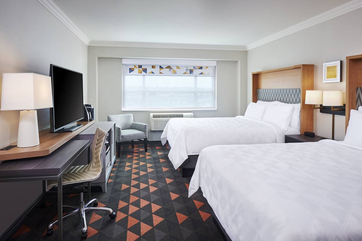 A hotel room with two beds, a desk with a computer, a chair, a TV, and a window with a geometric curtain.