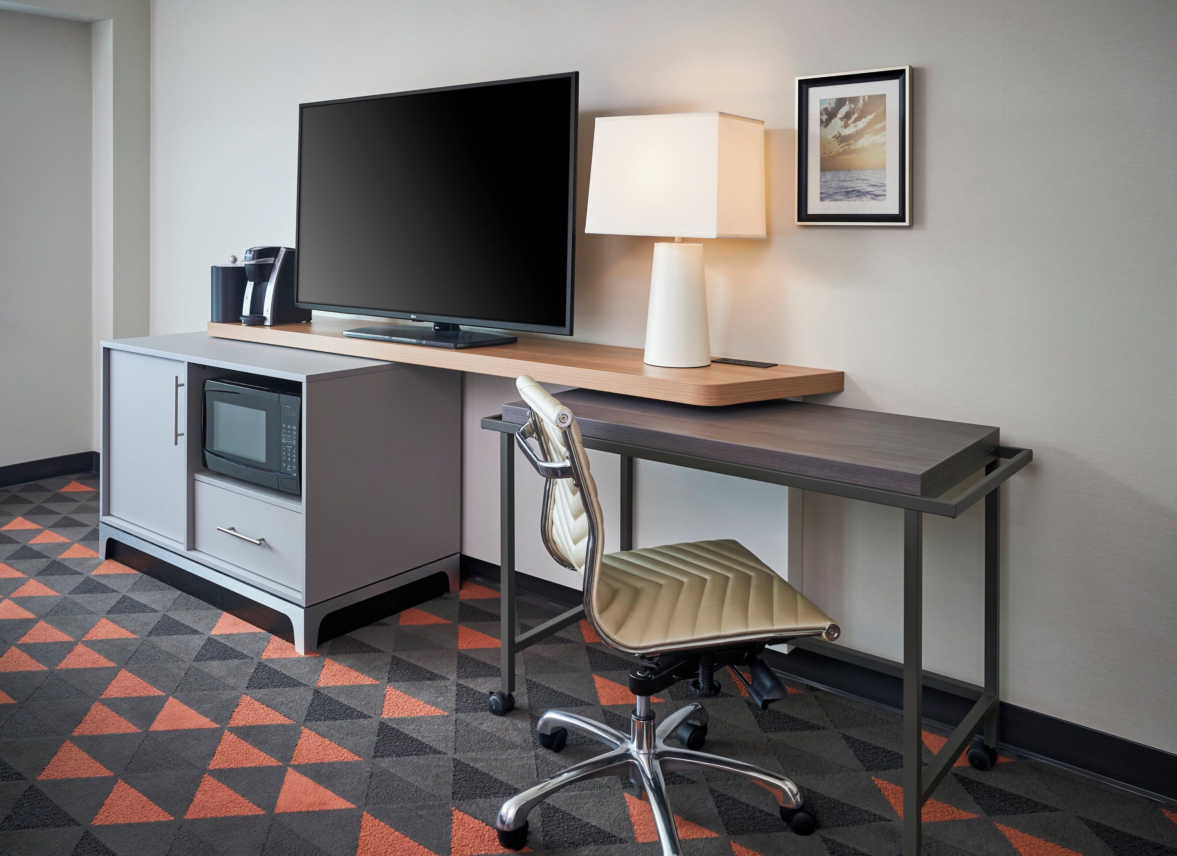 A hotel room with a desk, television, lamp, microwave, and a chair.