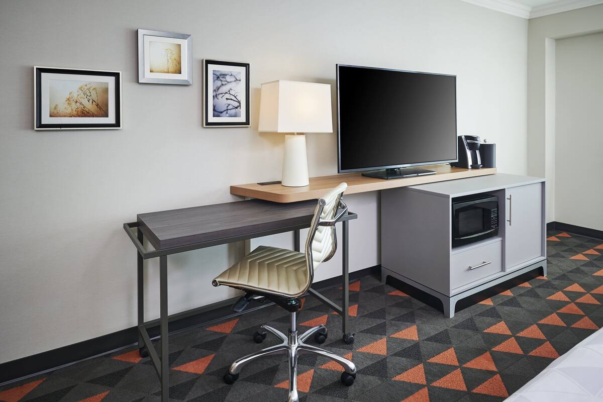 A hotel room with a desk, chair, television, microwave, and a cabinet.