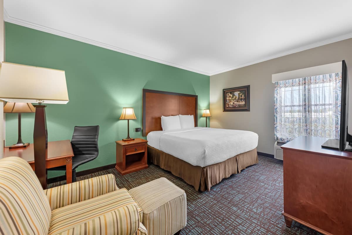 A hotel room with a bed, two bedside lamps, a chair, a desk, a dresser, and a window with curtains.
