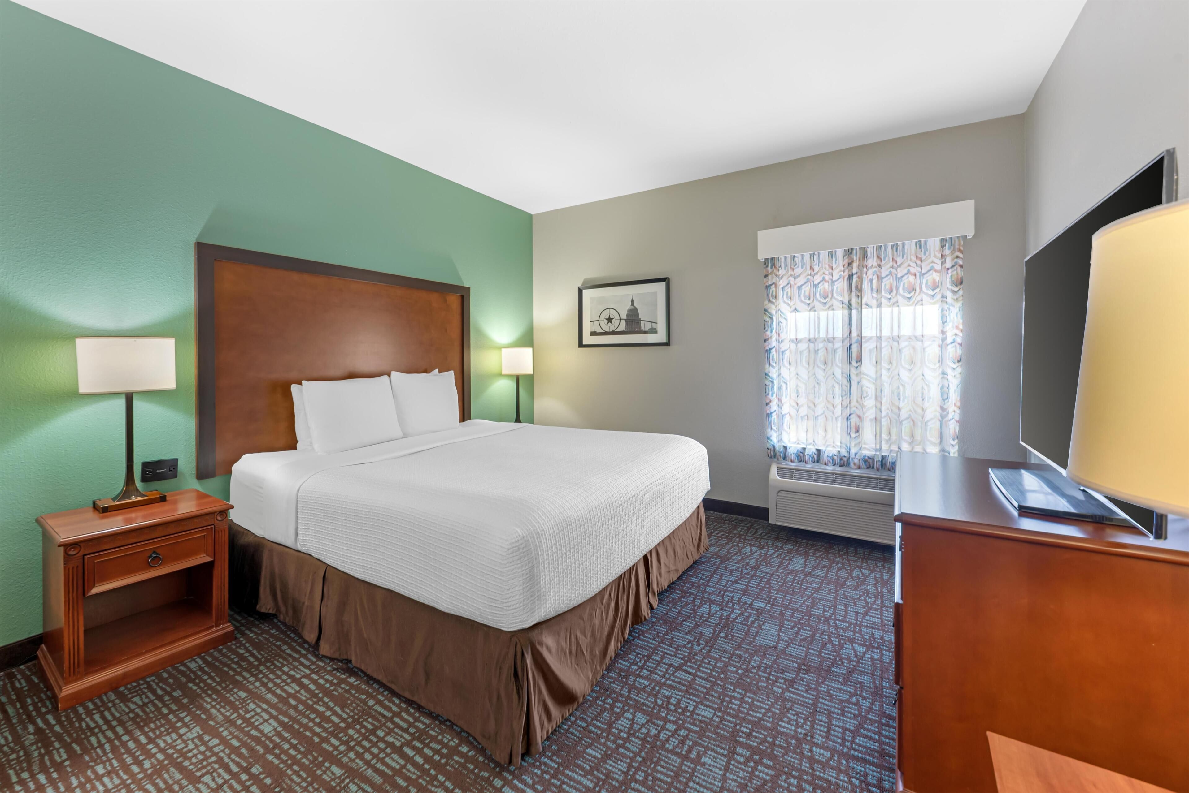 A hotel room with a bed, bedside tables, a lamp, a picture on the wall, a window with curtains, and a television.