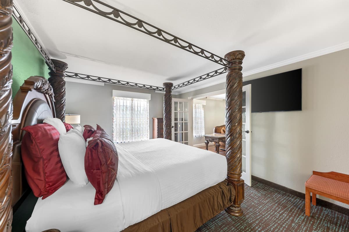 A four-poster bed with a canopy, a wooden bench, a flat-screen TV mounted on the wall, and a door leading to another room.