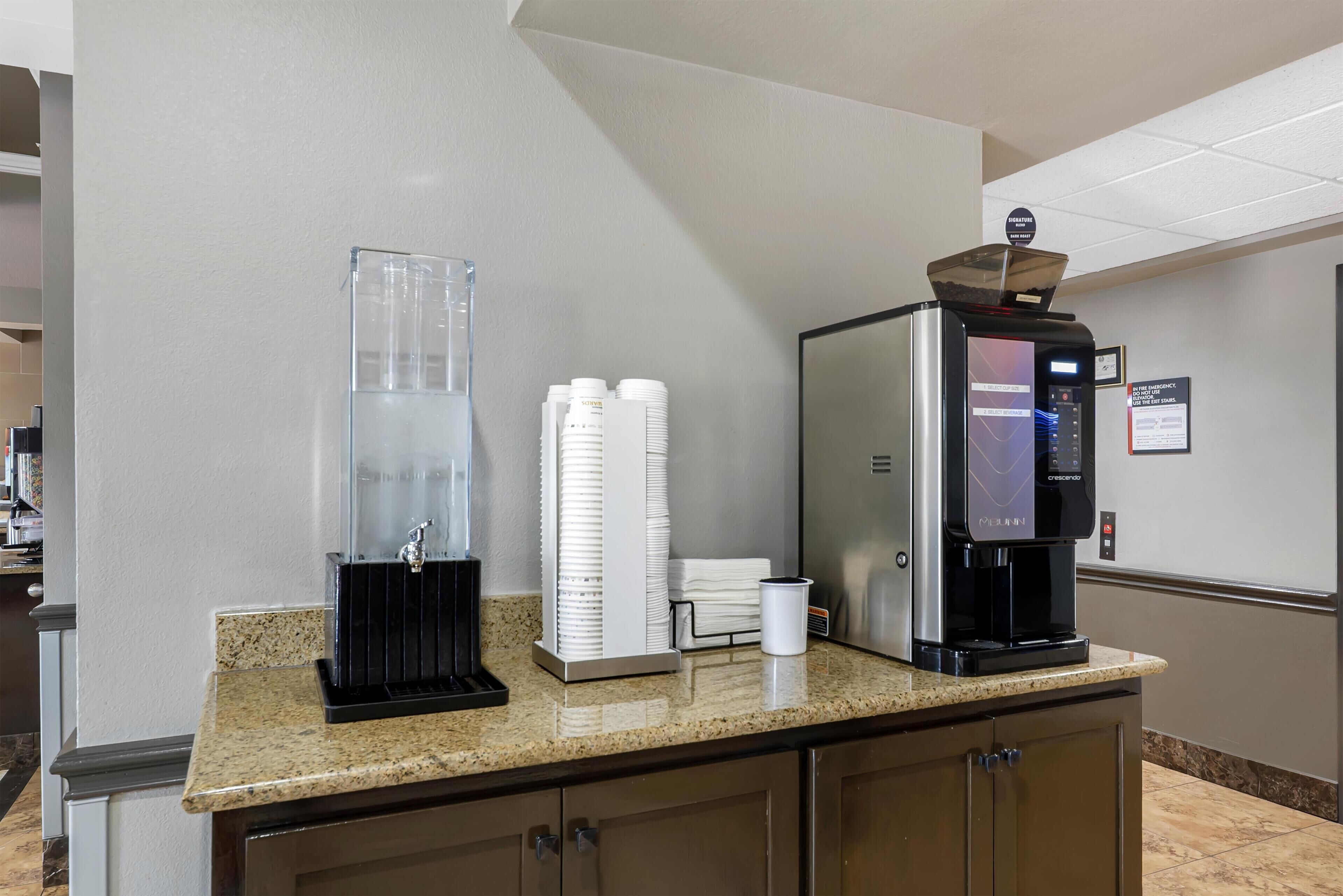 A self-service beverage station with a coffee machine, a water dispenser, and stacked paper cups.