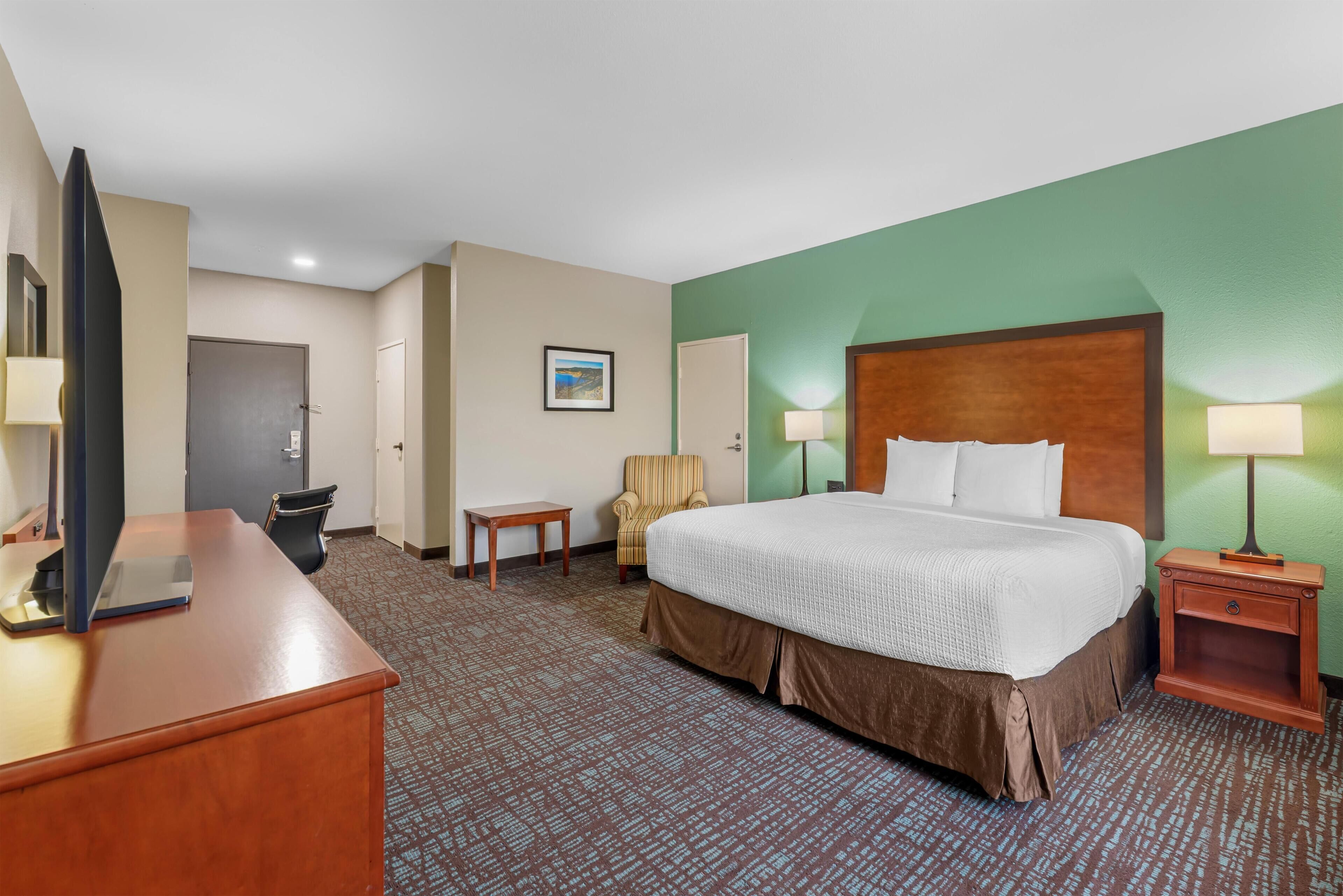 A hotel room with a large bed, a desk, a chair, a small table, a lamp, and a picture on the wall.