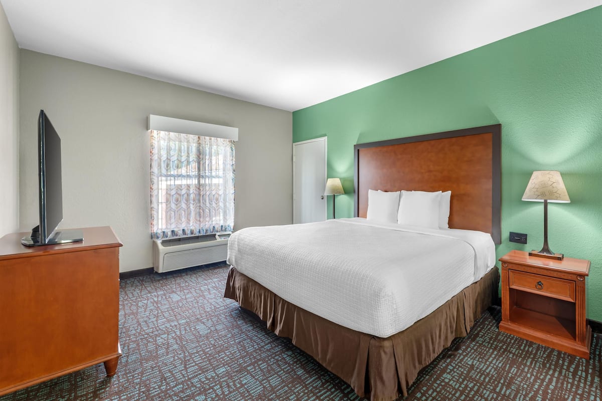 A hotel room with a large bed, a dresser, a television, a window with curtains, and a nightstand with a lamp.