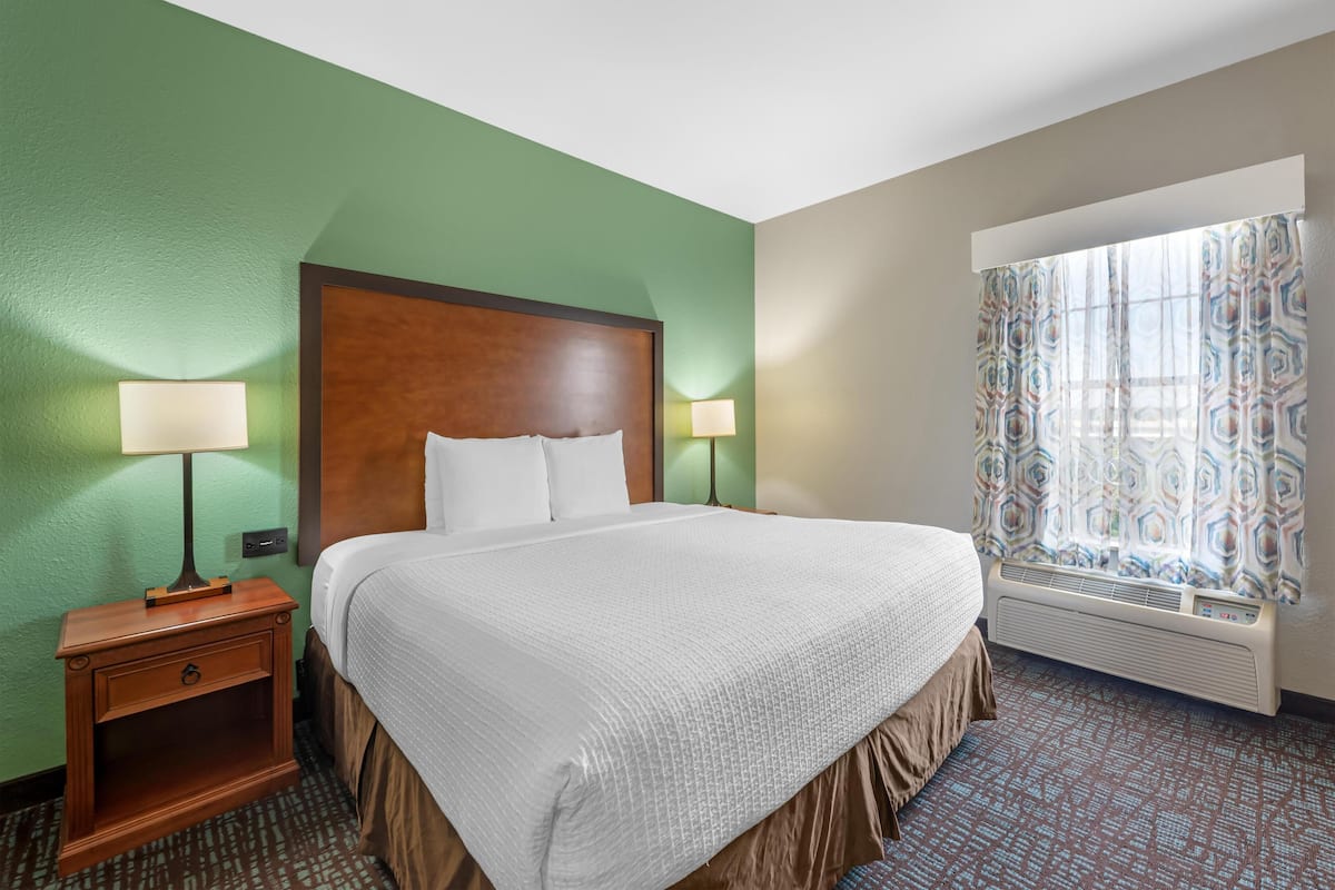 A hotel room with a bed, nightstand, lamp, window with curtains, and a wall heater.