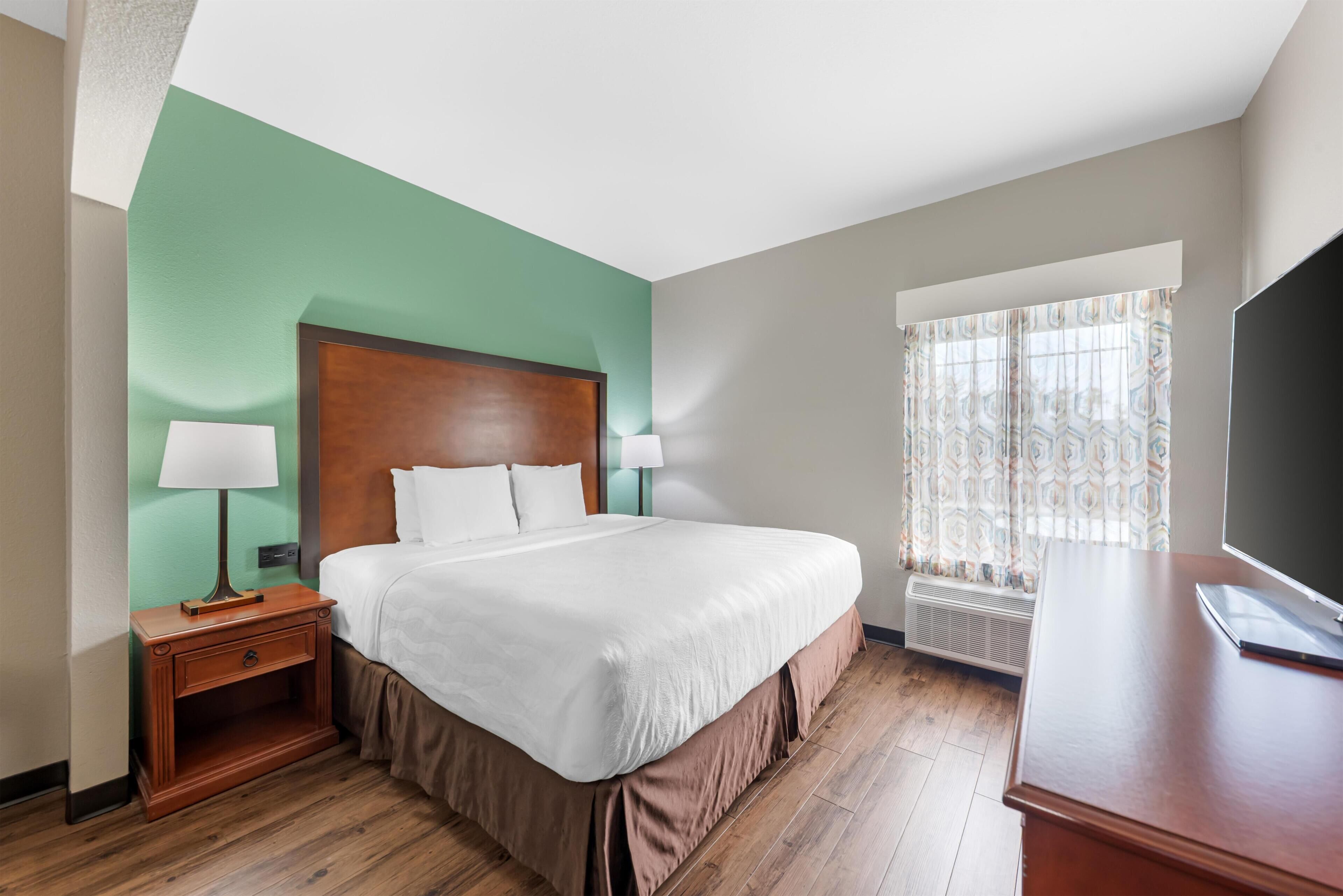 Una habitación de hotel con cama, mesita de noche, lámpara, televisor y ventana con cortinas.