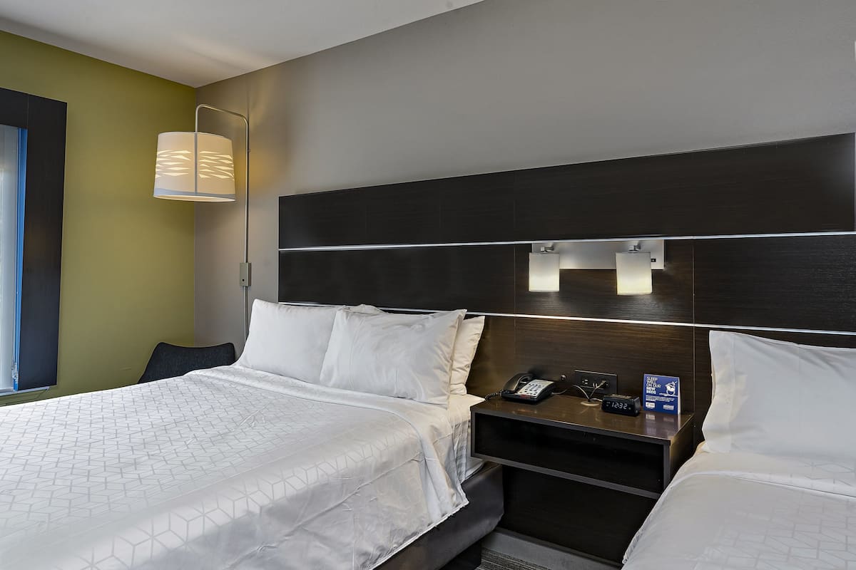 A hotel room with two beds, a bedside table with a phone and a clock, and a wall-mounted lamp.