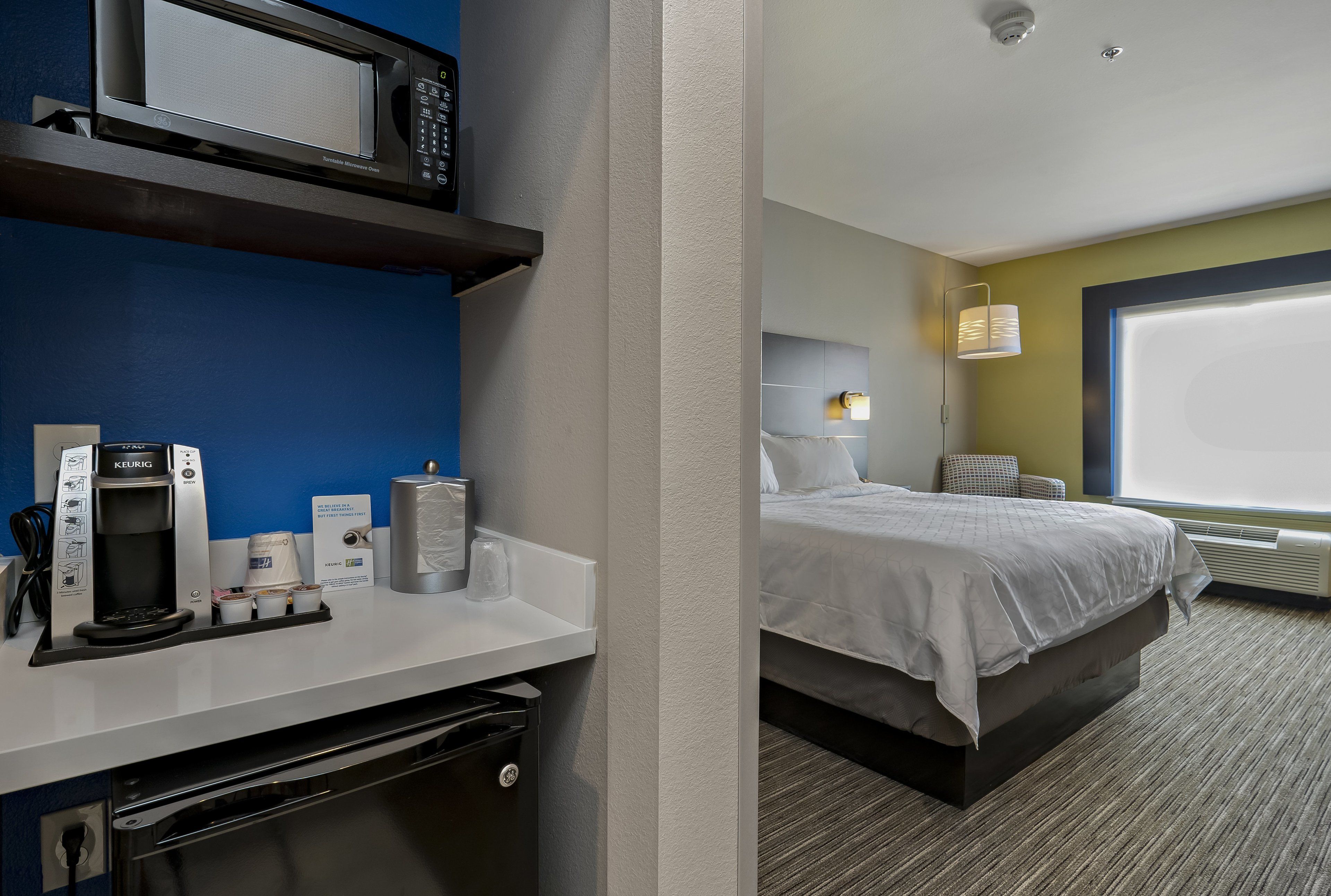 A hotel room with a bed, a desk with a coffee maker, a microwave, and a window with blinds.