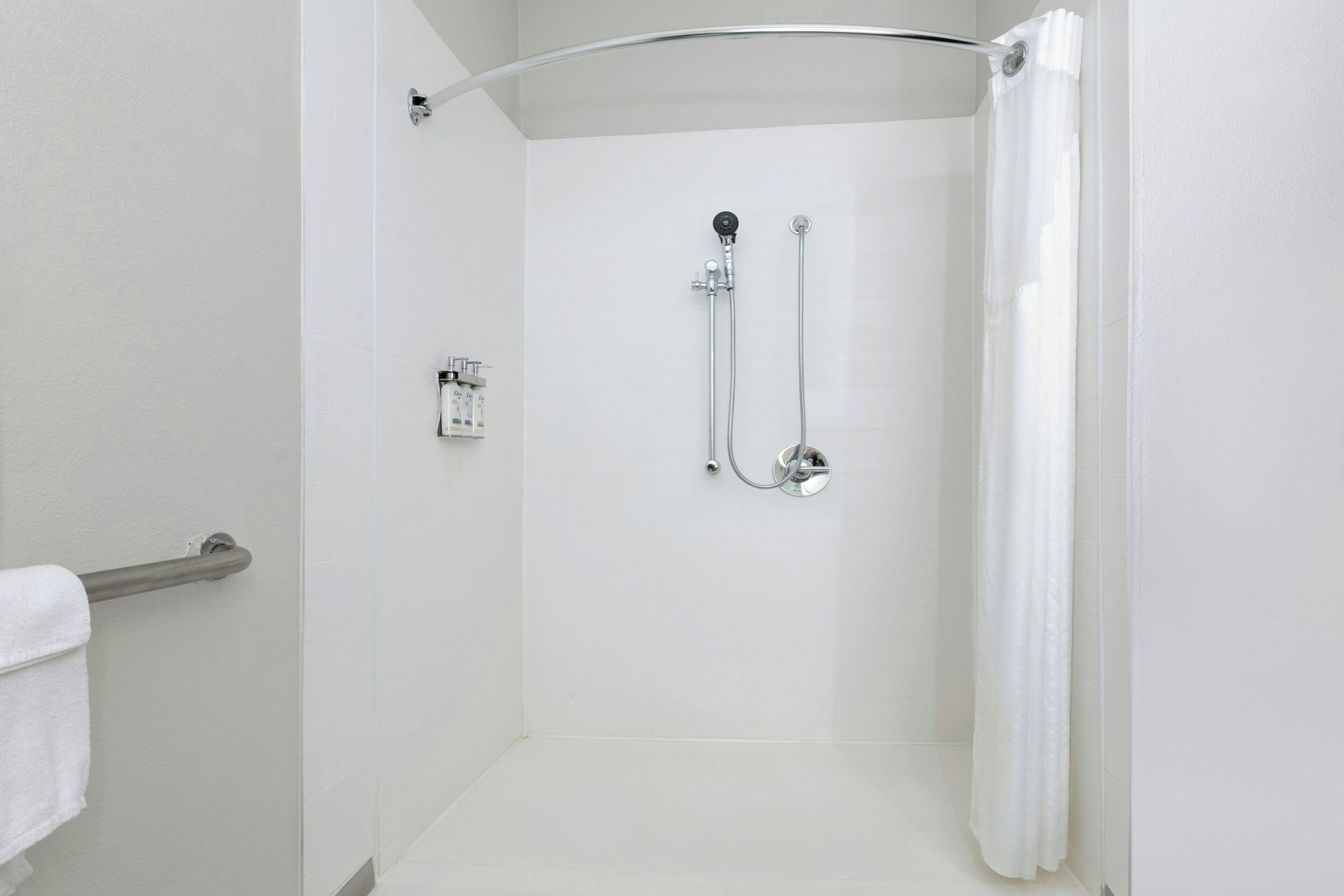 Baño blanco con puerta de vidrio, cabezal de ducha de mano, grifo empotrado en la pared y un perchero.