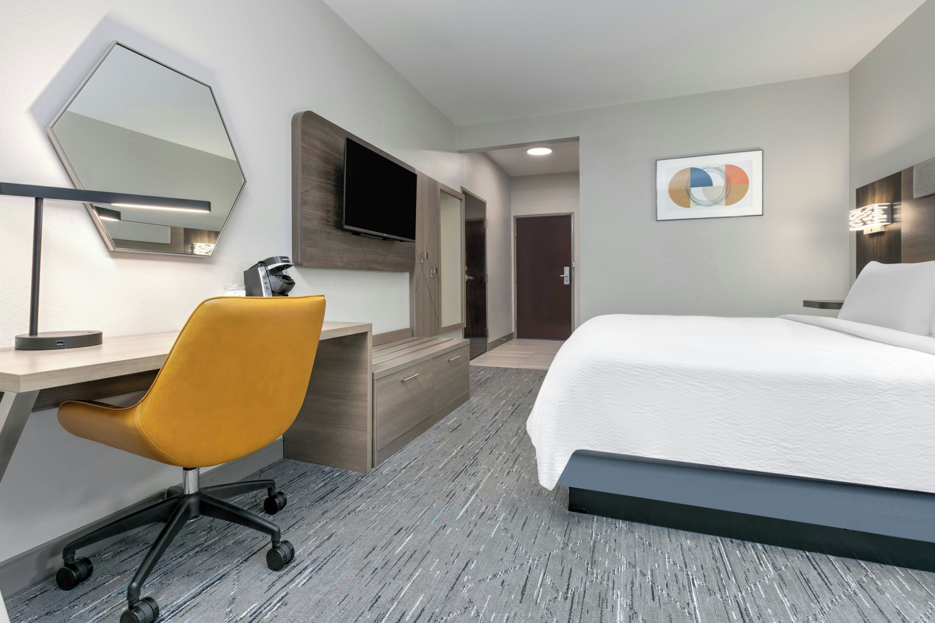 Habitación de hotel con cama, escritorio con silla, televisión de pantalla plana, aparador de madera, puerta y una obra de arte colgada en la pared.