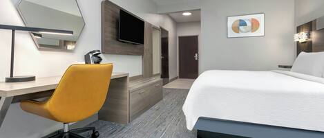 Habitación de hotel con cama, escritorio con silla, televisión de pantalla plana, aparador de madera, puerta y una obra de arte colgada en la pared.