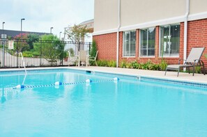 Una piscina con superficie de agua azul cristalina, rodeada por un edificio de ladrillo con grandes ventanales y una valla de metal negro.