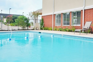 Piscina com superfície de água azul e límpida, rodeada por um edifício de tijolo com grandes janelas e uma cerca de metal preto.