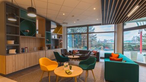 Ein modernes Interieur mit einem Holzbüro, grünen und orangefarbenen Sesseln, einem runden Tisch und einem petrolfarbenen Sofa mit orangefarbenen Kissen.