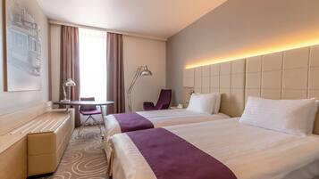 A hotel room with two beds, a desk, a chair, a lamp, and a view of a cityscape.