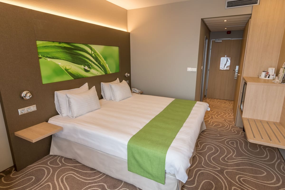 A hotel room with a large bed, a bedside table, a green throw, a wall-mounted TV, and a wooden cabinet with a mirror.
