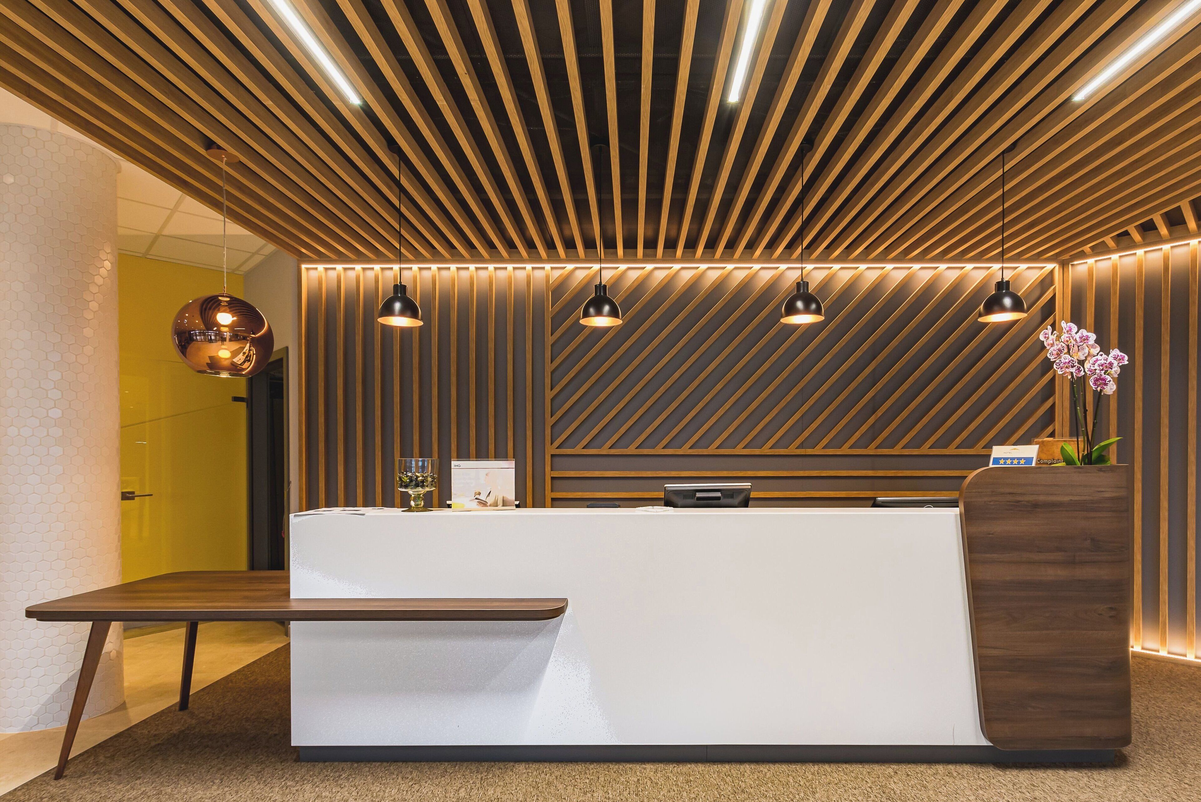 A reception desk with a modern design, wooden slats on the ceiling, pendant lighting, and a vase with flowers.