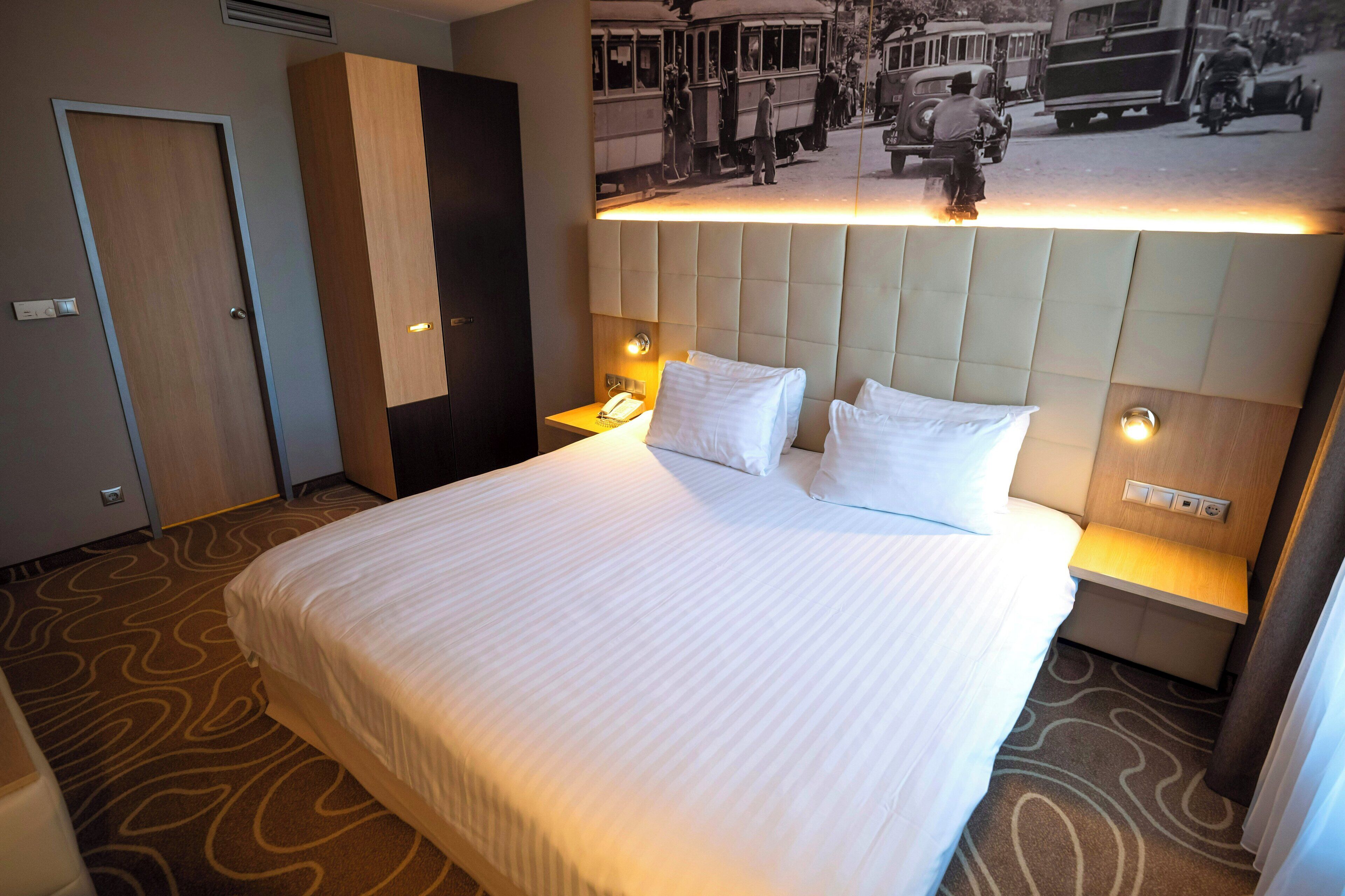 Habitació d'hotel amb un llit gran, un reposacapses amb un telèfon integrat i un quadre penjat a la paret que mostra una escena de carrer antiga.