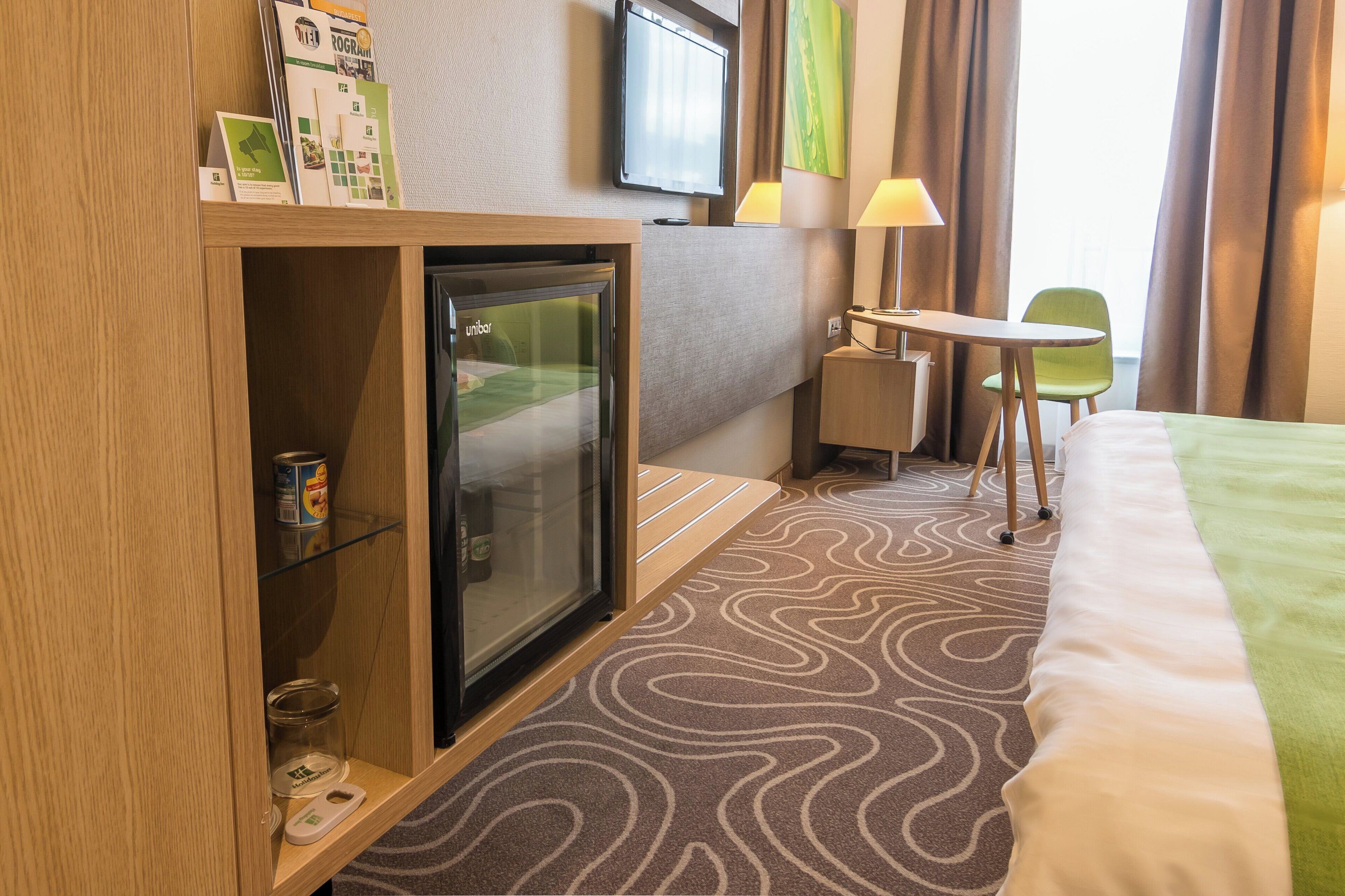 A hotel room with a bed, a desk with a chair, a minibar, a television, and a window with curtains.