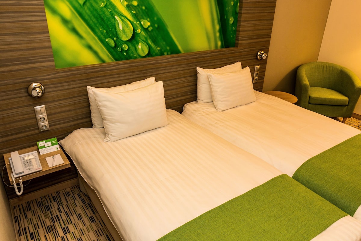 A hotel room with two beds, a green armchair, a telephone, and a wall-mounted artwork.