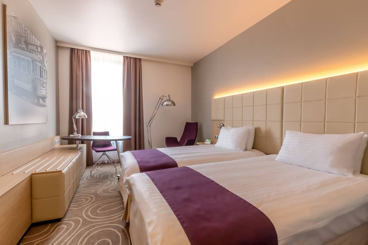 A hotel room with two beds, a desk, a chair, and a large window with curtains.