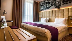 Ein Hotelzimmer mit einem großen Bett, einem Nachttisch aus Holz, einem Schreibtisch und einem Fenster mit Vorhängen.