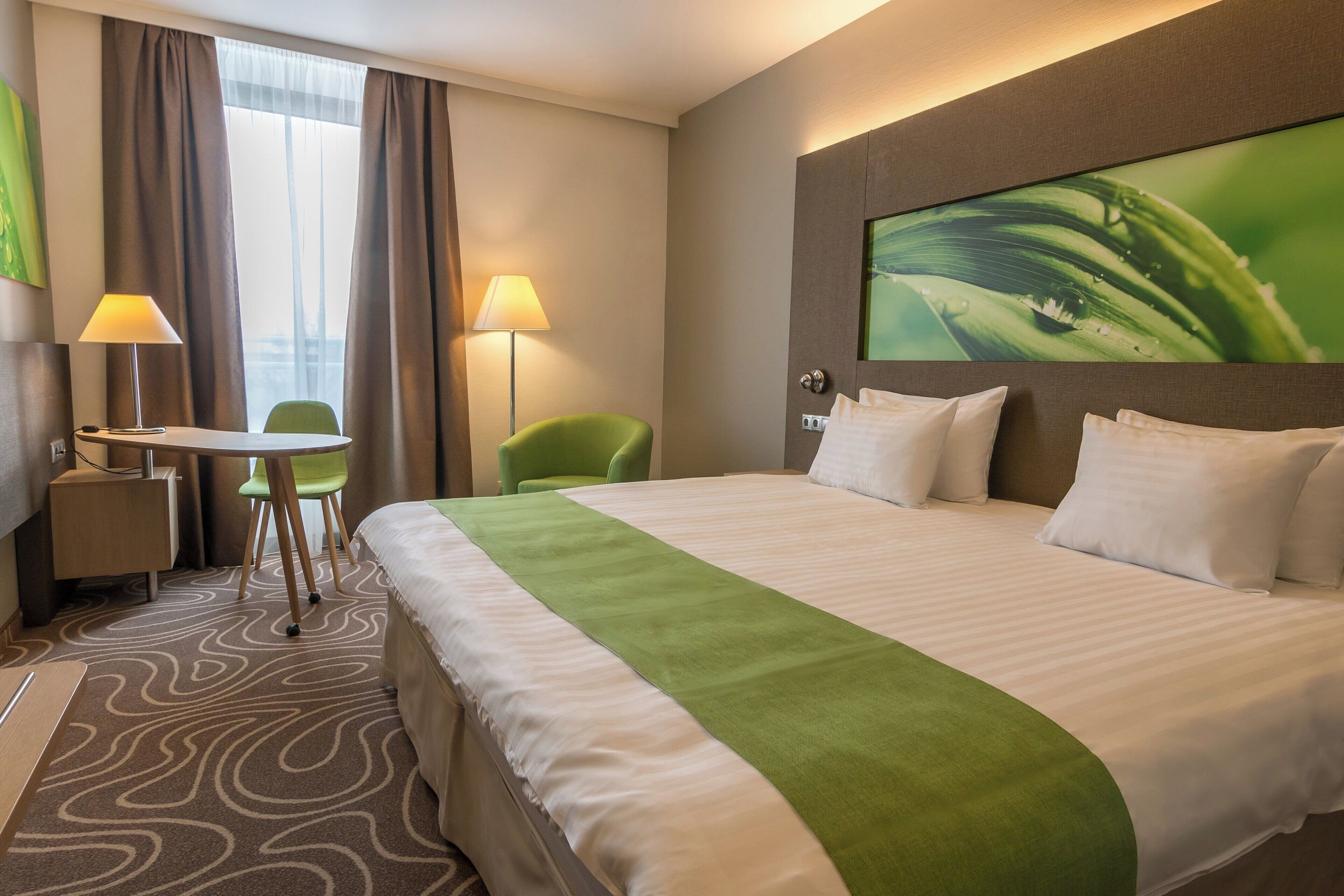 A hotel room with a large bed, a desk, two chairs, a lamp, and a painting on the wall.