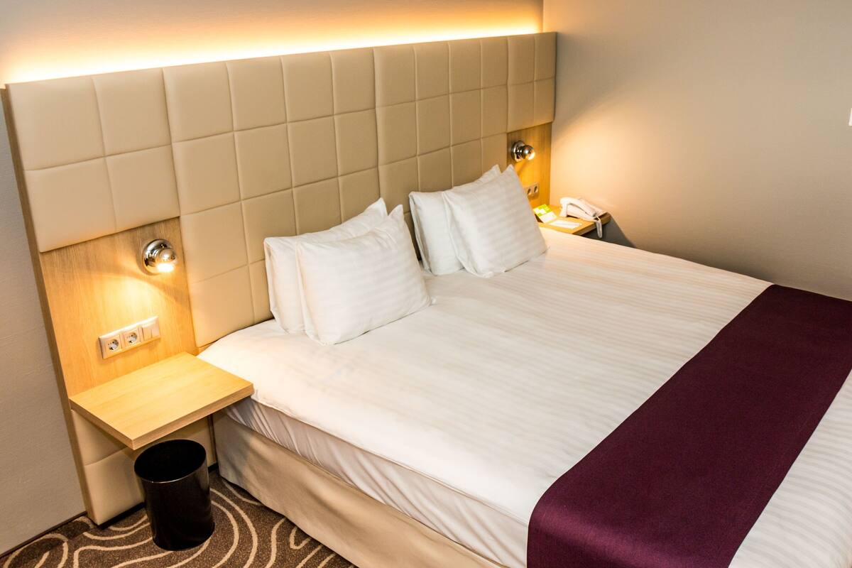 A hotel room with a large bed, a bedside table with a lamp, a trash can, and a wall socket.