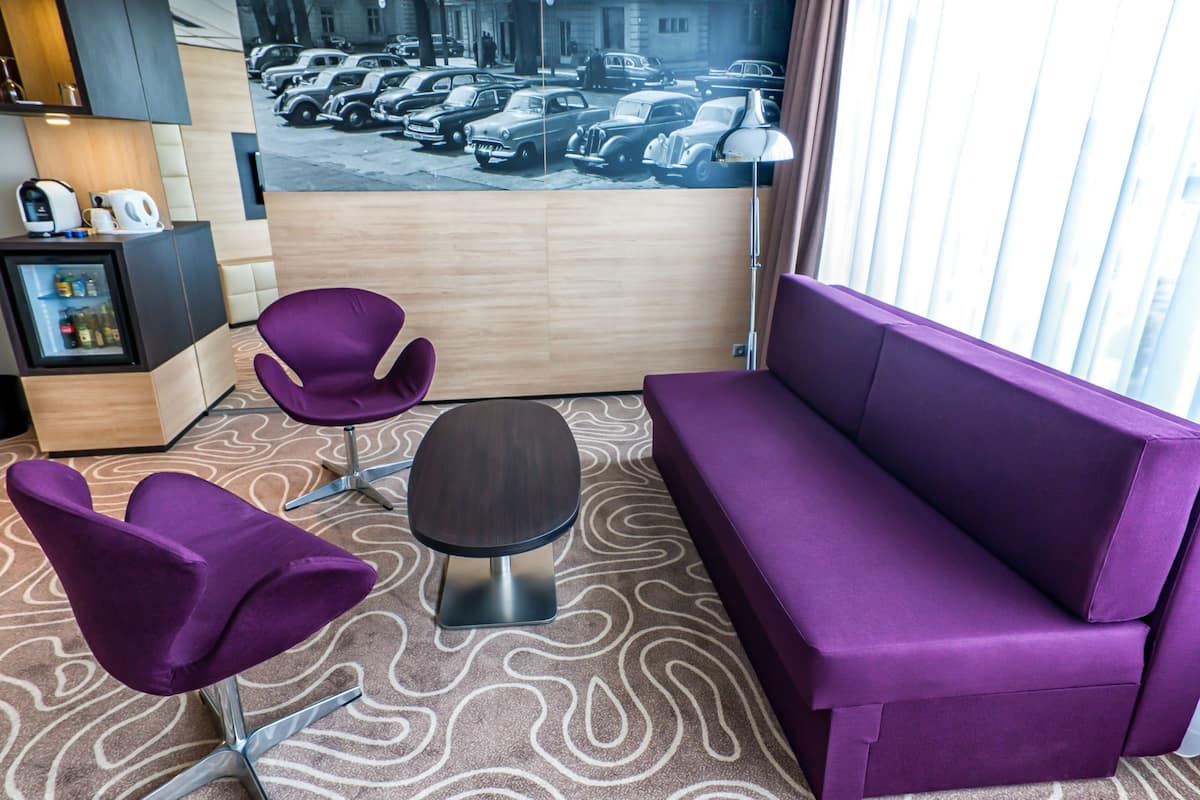 A modern hotel room with a purple sofa, two purple chairs, a small table, a minibar, and a large window with a view of parked cars.