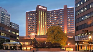 Un hotel de varios pisos con un llamativo letrero de "Hilton", rodeado de otros edificios y un árbol en primer plano.