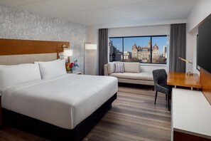 A hotel room with a large bed, a desk, a chair, a sofa, and a view of a cityscape through the window.
