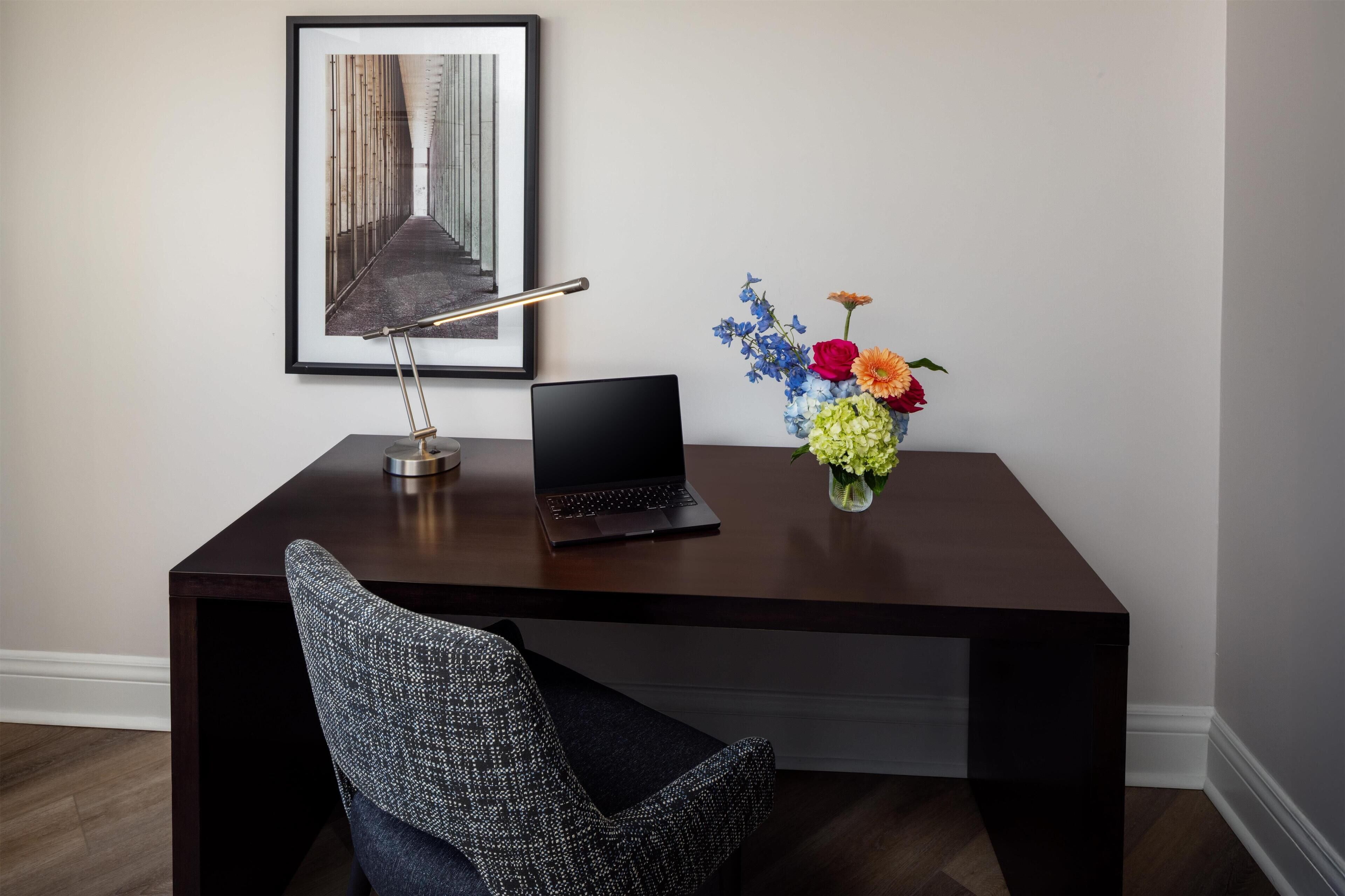 A dark wooden desk with a laptop, a framed photograph on the wall, and a vase with flowers.