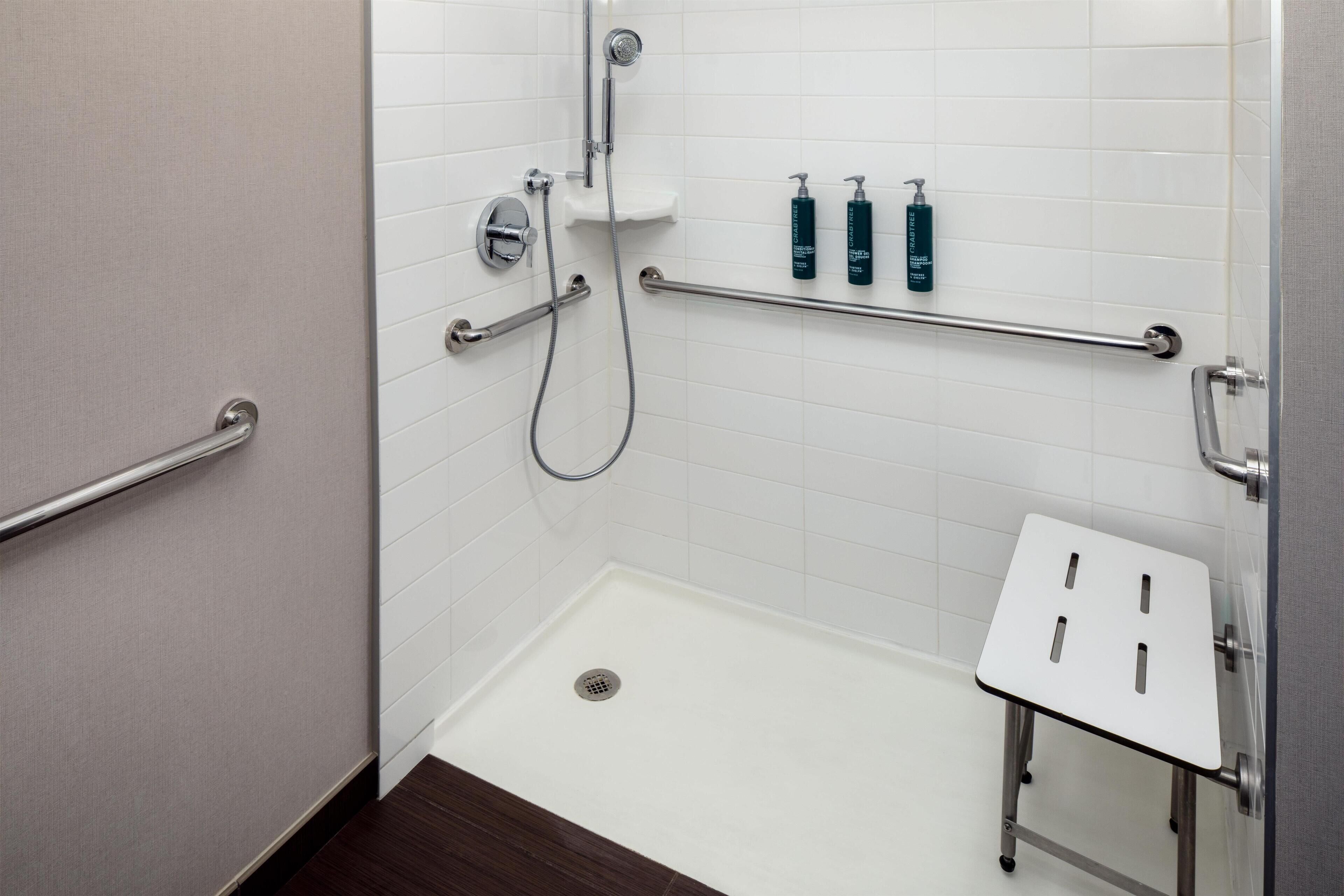 A bathroom with white tiled walls, a shower with a handheld showerhead, a grab bar, and a small white sink with a stool.