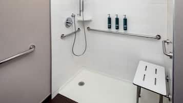 Un baño con paredes de azulejos blancos, una ducha con cabezal de ducha manual, una barra de apoyo y un lavamanos pequeño y blanco con un taburete.