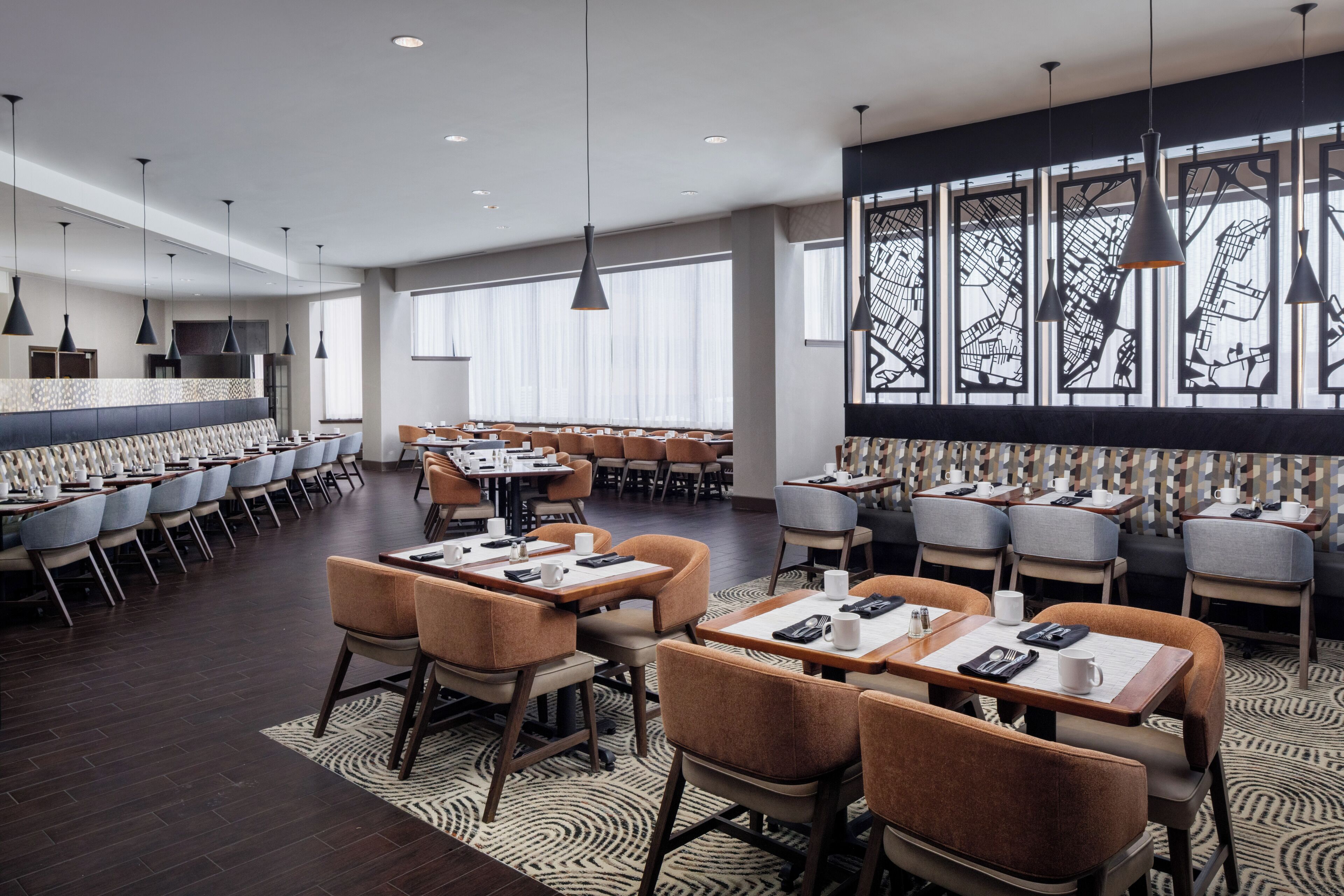 A modern hotel dining area with multiple tables and chairs, large windows with decorative black frames, and pendant lighting.