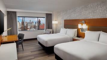 A hotel room with two beds, a desk, a chair, a lamp, and a view of a cityscape.