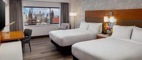A hotel room with two beds, a desk, a chair, a window with a city view, and a lamp.