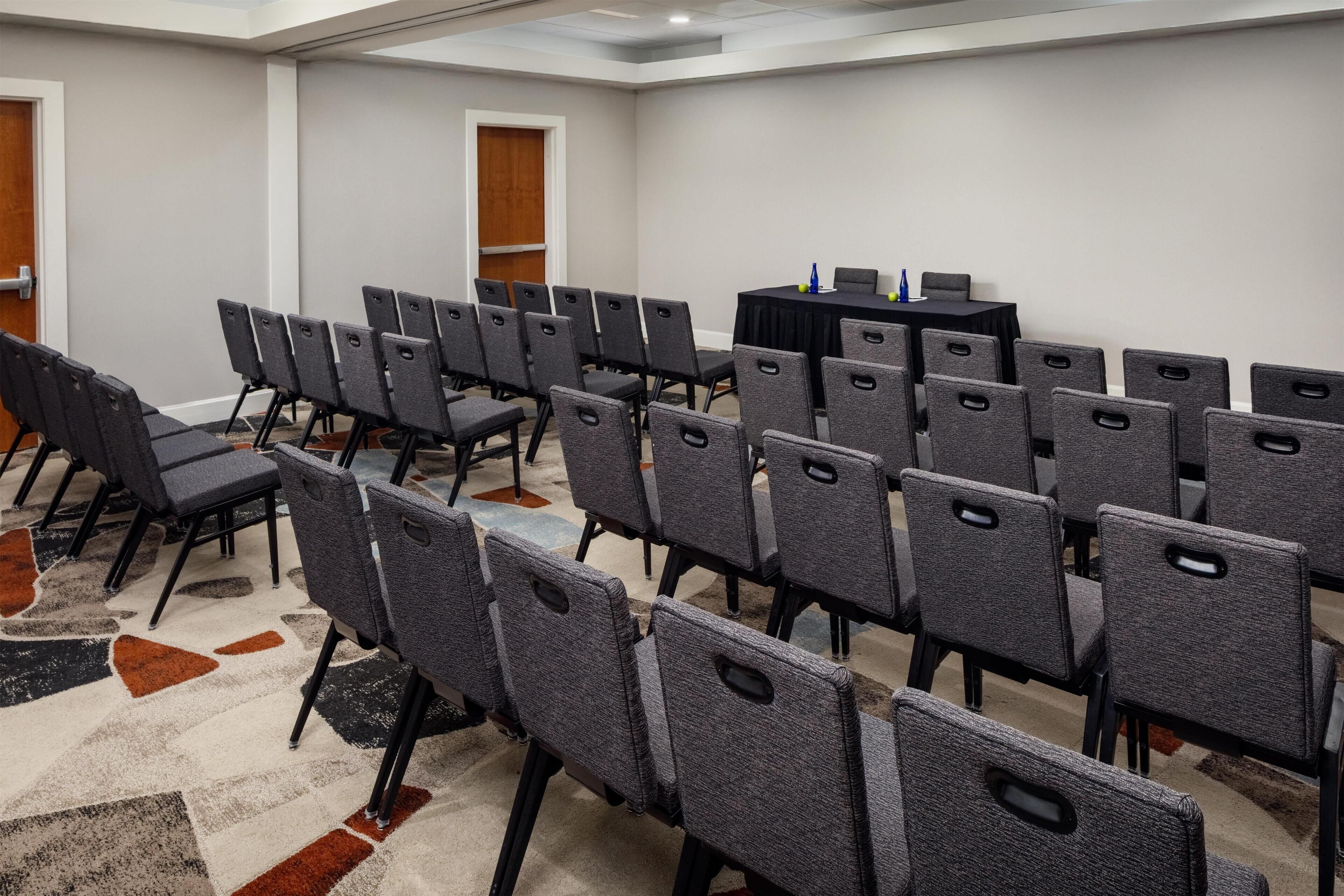 Uma sala de conferências com fileiras de cadeiras cinzas, uma mesa preta com garrafas de água azuis e um carpete com padrão geométrico.