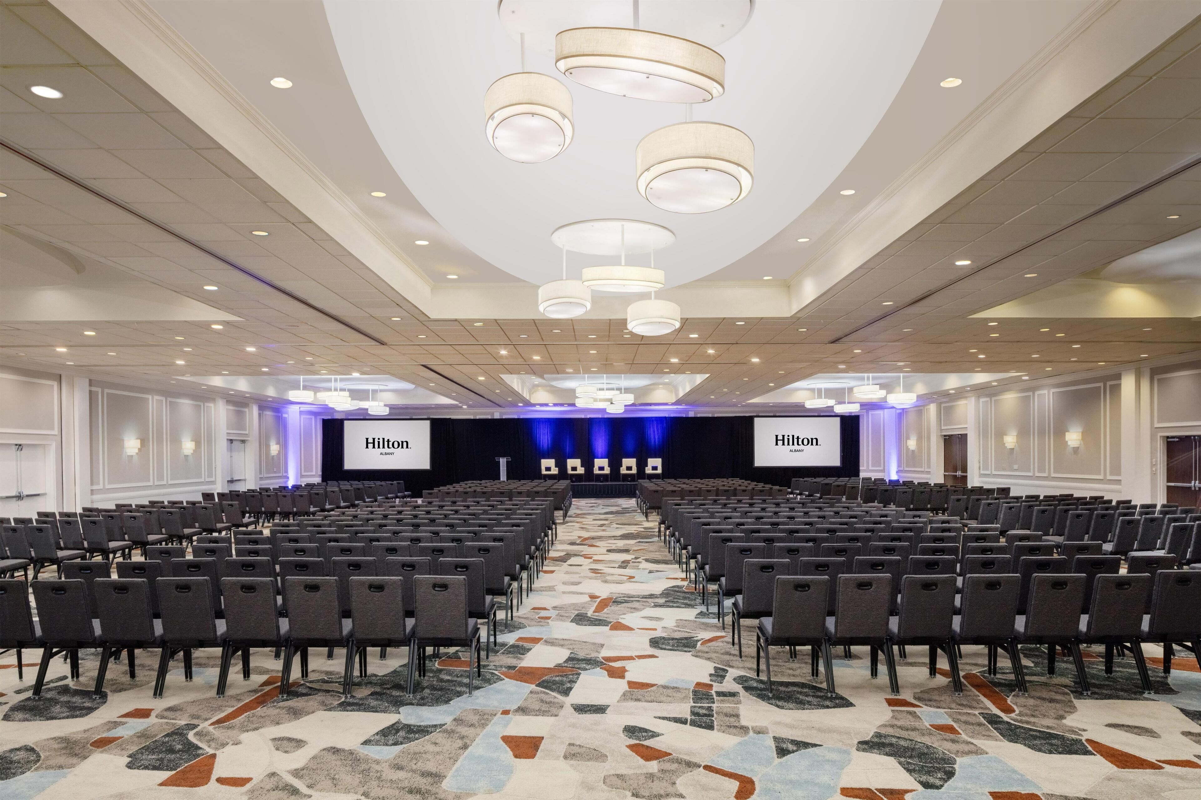 Un salón de conferencias de hotel con filas de sillas, un escenario con una pantalla 'Hilton' y una alfombra estampada.