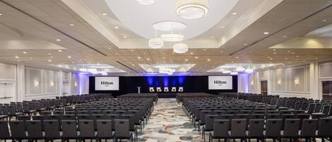Un salón de conferencias de hotel con filas de sillas, un escenario con una pantalla 'Hilton' y una alfombra estampada.
