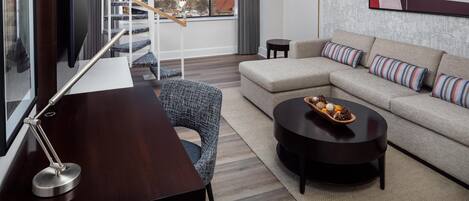 A modern living room with a sofa, coffee table, and a desk with a lamp. A large window offers a view of a cityscape and a building.
